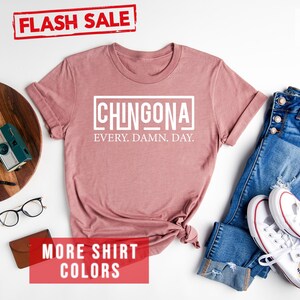 May include: A heather mauve t-shirt with the text "CHINGONA EVERY. DAMN. DAY." printed in white. The shirt is laid flat on a white background with a pair of jeans, white sneakers, and other accessories.