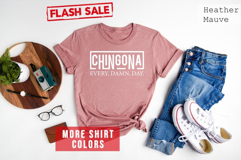 May include: A heather mauve t-shirt with the text "CHINGONA EVERY. DAMN. DAY." printed in white. The shirt is laid flat on a white background with a pair of jeans, white sneakers, and other accessories.