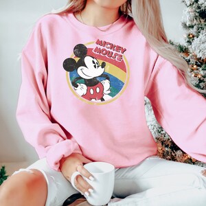 Puede incluir: Sudadera rosa con un gr&aacute;fico de Mickey Mouse de estilo vintage. El gr&aacute;fico presenta a Mickey Mouse con una camisa roja y pantalones cortos amarillos, de pie con las manos en las caderas. El gr&aacute;fico est&aacute; rodeado por un c&iacute;rculo de colores arco&iacute;ris con el texto "Mickey Mouse" en letras amarillas.