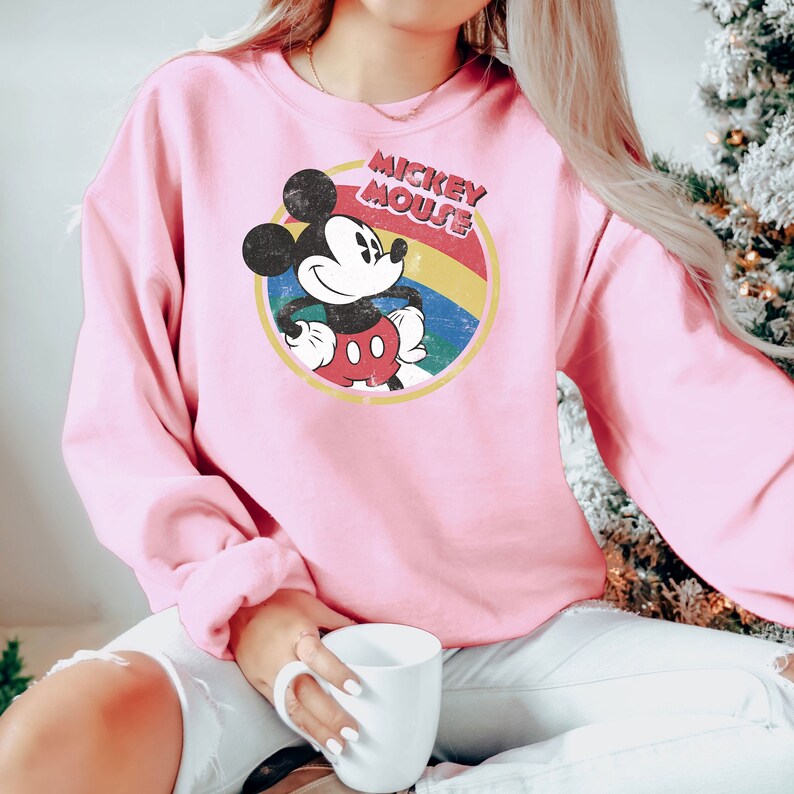 Puede incluir: Sudadera rosa con un gr&aacute;fico de Mickey Mouse de estilo vintage. El gr&aacute;fico presenta a Mickey Mouse con una camisa roja y pantalones cortos amarillos, de pie con las manos en las caderas. El gr&aacute;fico est&aacute; rodeado por un c&iacute;rculo de colores arco&iacute;ris con el texto "Mickey Mouse" en letras amarillas.