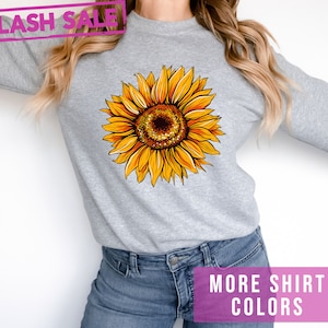 May include: A gray sweatshirt with a yellow sunflower graphic. The text "MORE SHIRT COLORS" is printed in pink at the bottom of the image.