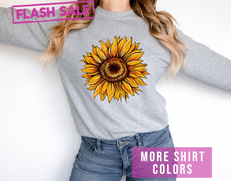 May include: A gray sweatshirt with a yellow sunflower graphic. The text "MORE SHIRT COLORS" is printed in pink at the bottom of the image.