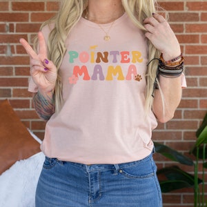 May include: A pink t-shirt with the text "POINTER MAMA" in colourful retro letters. The letters are outlined in black and have a flower and paw print design.