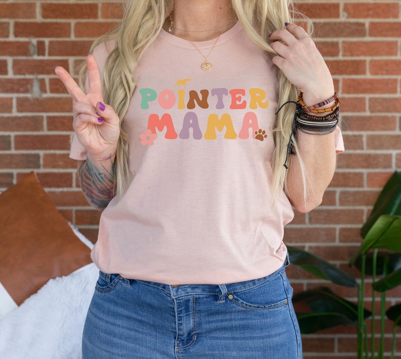 May include: A pink t-shirt with the text "POINTER MAMA" in colourful retro letters. The letters are outlined in black and have a flower and paw print design.