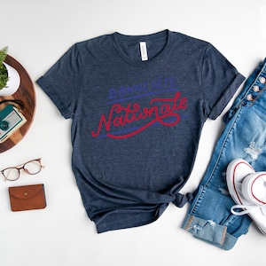 May include: A heather navy short-sleeve t-shirt featuring the text "Bonne F&ecirc;te Nationale" in blue and red script lettering. The shirt is displayed alongside blue denim jeans, white sneakers, a watch, glasses, a small wallet, and a plant.