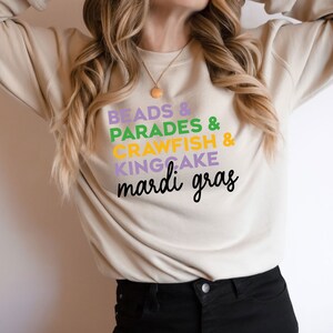May include: A beige sweatshirt with the text "Beads & Parades & Crawfish & Kingcake Mardi Gras" printed on the front in purple, green, yellow, and black.