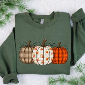 May include: Green sweatshirt featuring three decorative pumpkins. The pumpkins are brown plaid, white floral, and orange plaid. The pumpkins are connected by a black stitched line. The sweatshirt has long sleeves.