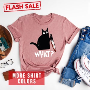 Op de afbeelding: Een heide-paarse t-shirt met een zwarte kat die een bloedig mes vasthoudt en de tekst "WHAT?" erop gedrukt. Het shirt ligt plat op een witte achtergrond met jeans, witte sneakers en andere accessoires.