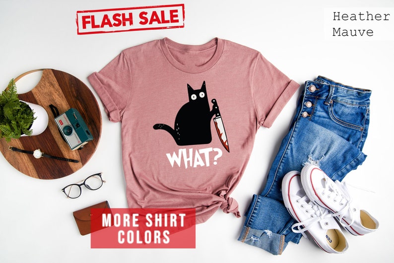 Op de afbeelding: Een heide-paarse t-shirt met een zwarte kat die een bloedig mes vasthoudt en de tekst "WHAT?" erop gedrukt. Het shirt ligt plat op een witte achtergrond met jeans, witte sneakers en andere accessoires.