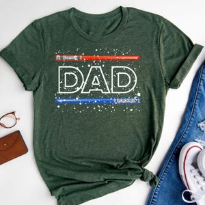 May include: Dark green t-shirt with the word "DAD" in white, stylized text. Above and below the word are red and blue lightsaber designs. White speckles are scattered around the text and lightsabers. The shirt is folded on a white surface.
