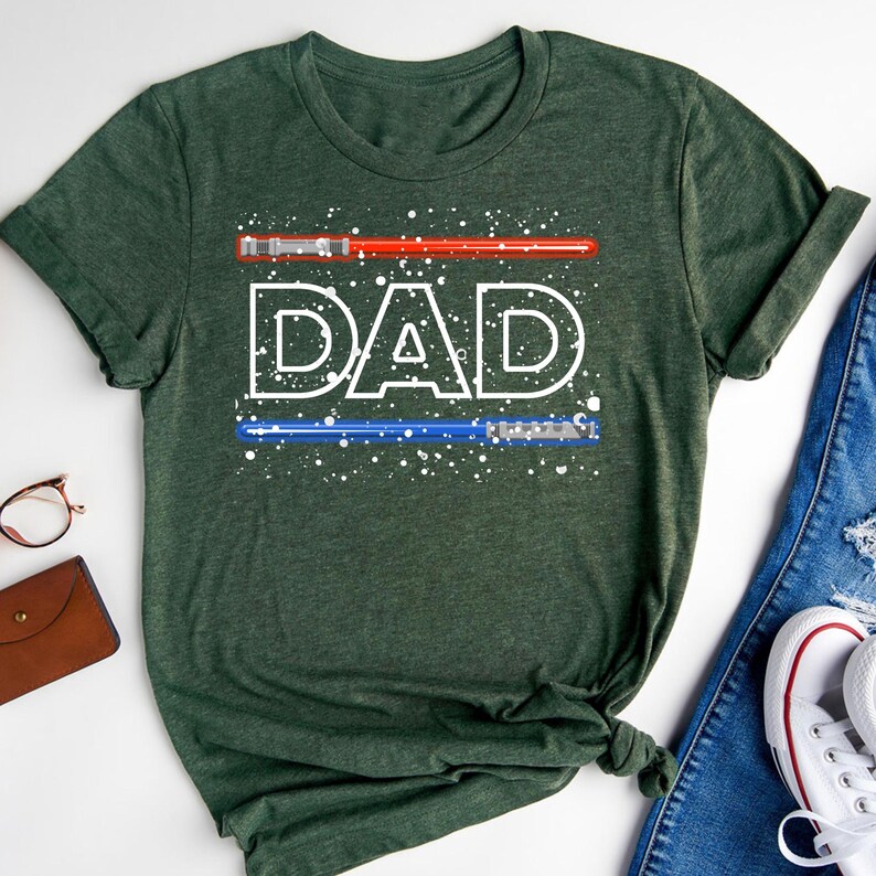 May include: Dark green t-shirt with the word "DAD" in white, stylized text. Above and below the word are red and blue lightsaber designs. White speckles are scattered around the text and lightsabers. The shirt is folded on a white surface.