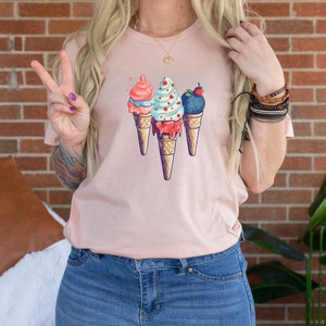 May include: A pink t-shirt with a graphic of three ice cream cones. The ice cream cones are decorated with different flavors and toppings, including a red and white swirl, a blue and white swirl, and a dark blue swirl with a red cherry on top.