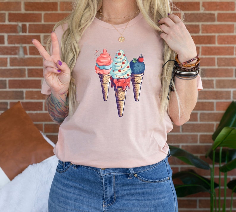 May include: A pink t-shirt with a graphic of three ice cream cones. The ice cream cones are decorated with different flavors and toppings, including a red and white swirl, a blue and white swirl, and a dark blue swirl with a red cherry on top.