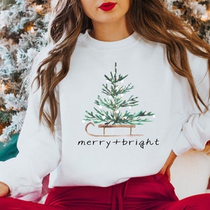 May include: White sweatshirt with a watercolor Christmas tree design on a sled, with the words "merry + bright" in a handwritten font. The sweatshirt is paired with red pants.
