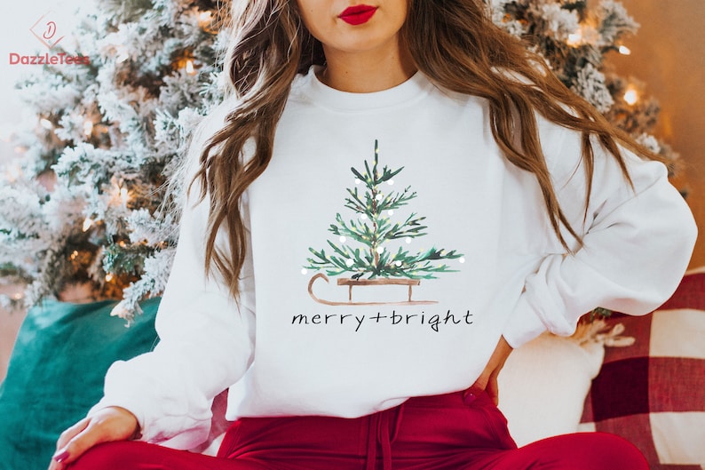 May include: White sweatshirt with a watercolor Christmas tree design on a sled, with the words "merry + bright" in a handwritten font. The sweatshirt is paired with red pants.