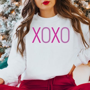 May include: White sweatshirt with a pink XOXO graphic print. The sweatshirt is being worn by a person sitting in front of a Christmas tree.