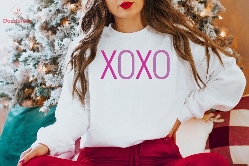May include: White sweatshirt with a pink XOXO graphic print. The sweatshirt is being worn by a person sitting in front of a Christmas tree.