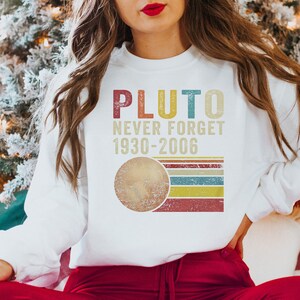 Op de afbeelding: Wit sweatshirt met een vintage-stijl grafische afbeelding met de tekst "Pluto Never Forget 1930-2006". De afbeelding toont een gestileerde afbeelding van Pluto met een regenboogstreep achtergrond.