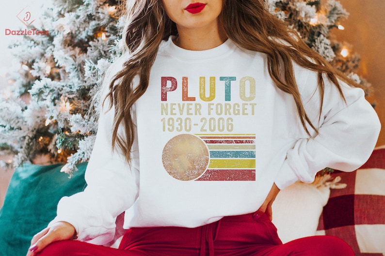 Op de afbeelding: Wit sweatshirt met een vintage-stijl grafische afbeelding met de tekst "Pluto Never Forget 1930-2006". De afbeelding toont een gestileerde afbeelding van Pluto met een regenboogstreep achtergrond.