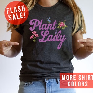 May include: A dark gray t-shirt with the text "Plant Lady" in purple script with pink flowers. The text "More Shirt Colors" is in red at the bottom of the image.