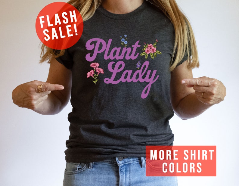 May include: A dark gray t-shirt with the text "Plant Lady" in purple script with pink flowers. The text "More Shirt Colors" is in red at the bottom of the image.