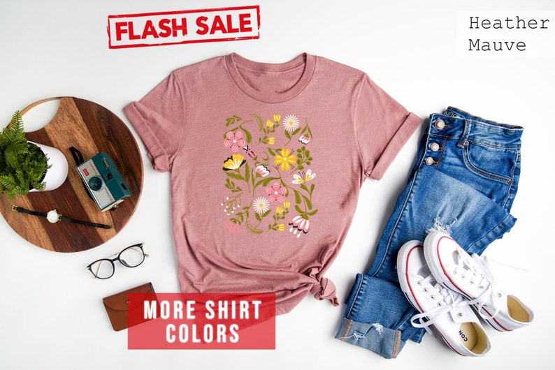 May include: A heather mauve t-shirt with a floral print featuring butterflies and flowers in pink, yellow, and green. The text "FLASH SALE" and "MORE SHIRT COLORS" is visible in red. The shirt is displayed on a white background with a pair of glasses, a wooden tray, a camera, and a pair of blue jeans with white sneakers.