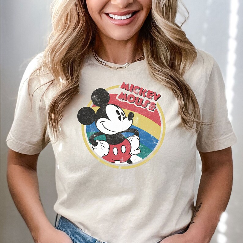 Puede incluir: Una camiseta beige con un gr&aacute;fico de estilo vintage de Mickey Mouse. El gr&aacute;fico presenta a Mickey Mouse con un atuendo rojo y negro, un c&iacute;rculo amarillo con un arco&iacute;ris y el texto "Mickey Mouse".