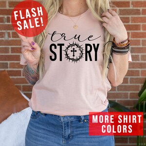 May include: A pink t-shirt with the text "true STORY" in black lettering. The "O" in "STORY" is replaced with a crown of thorns with a cross in the center. The shirt is being modeled by a person wearing blue jeans.