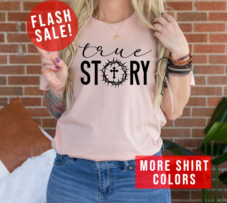 May include: A pink t-shirt with the text "true STORY" in black lettering. The "O" in "STORY" is replaced with a crown of thorns with a cross in the center. The shirt is being modeled by a person wearing blue jeans.