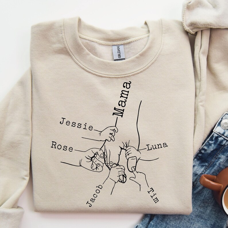 May include: A beige sweatshirt with a black line drawing of hands holding onto a central figure labeled "Mama". The hands are labeled with the names "Jessie", "Rose", "Jacob", "Tim", and "Luna".