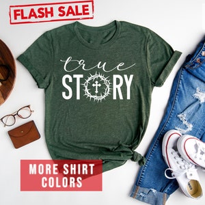 May include: A forest green t-shirt with the words "true story" in white text. The word "story" is surrounded by a white crown of thorns with a cross in the center. The shirt is laid flat on a white surface with a pair of jeans, white sneakers, and other accessories.