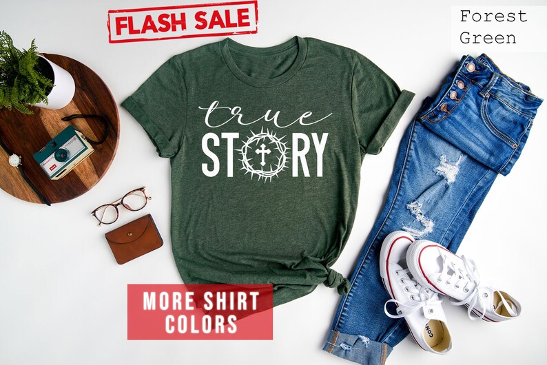 May include: A forest green t-shirt with the words "true story" in white text. The word "story" is surrounded by a white crown of thorns with a cross in the center. The shirt is laid flat on a white surface with a pair of jeans, white sneakers, and other accessories.