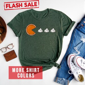 K&ouml;nnte beinhalten: Ein T-Shirt in Waldgr&uuml;n mit einem wei&szlig;en Pac-Man-Design, das drei wei&szlig;e Mini-Marshmallows frisst. Der Text "FLASH SALE" ist oben im Bild in Rot. Der Text "MORE SHIRT COLORS" ist unten im Bild in Rot.