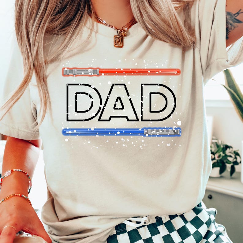 May include: Beige t-shirt with the word "DAD" in black outlined letters. Above and below the word are red and blue lightsaber graphics. White speckles are scattered across the design. The shirt is a casual style.