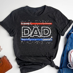 May include: Dark gray t-shirt with the word "DAD" in white, with a red lightsaber above and a blue lightsaber below. White speckles are scattered around the text and lightsabers. The shirt is folded and tied at the bottom.