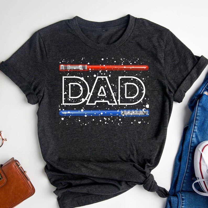 May include: Dark gray t-shirt with the word "DAD" in white, with a red lightsaber above and a blue lightsaber below. White speckles are scattered around the text and lightsabers. The shirt is folded and tied at the bottom.