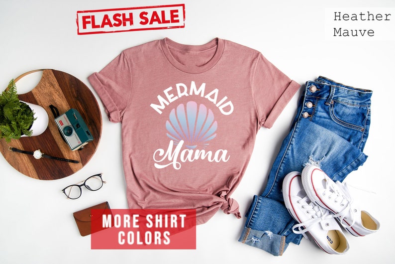 May include: A heather mauve t-shirt with the text "Mermaid Mama" in white. A seashell with a pink and blue ombre design is above the text. The shirt is laid flat on a white background with jeans, sneakers, and other accessories.