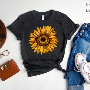 May include: A dark grey heather t-shirt with a yellow sunflower graphic. The shirt is laid flat on a white surface with a pair of jeans, a pair of white sneakers, a brown purse, and a camera.