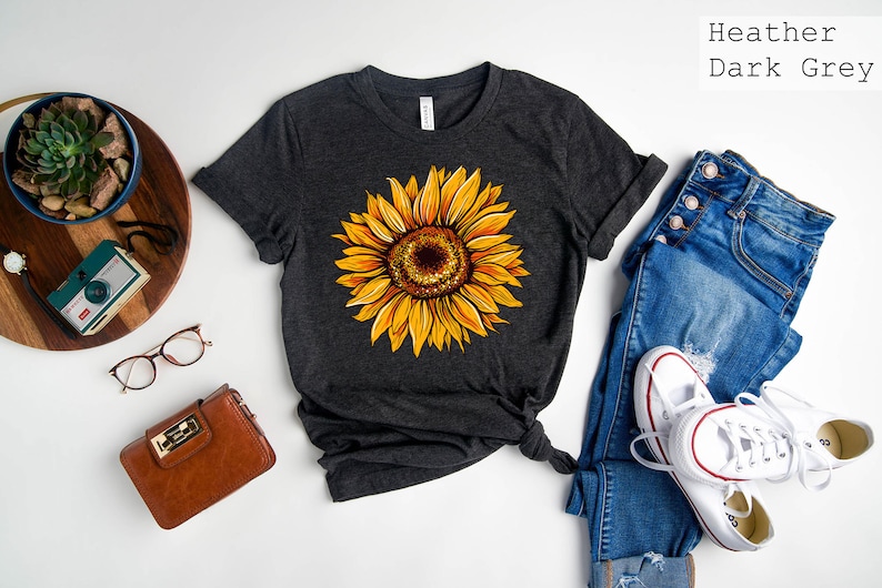 May include: A dark grey heather t-shirt with a yellow sunflower graphic. The shirt is laid flat on a white surface with a pair of jeans, a pair of white sneakers, a brown purse, and a camera.