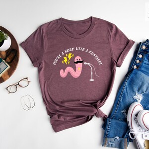 May include: A heather maroon t-shirt with a white graphic of a pink worm with a mustache holding a microphone. The text "You're a born with a mustache" is printed on the shirt.