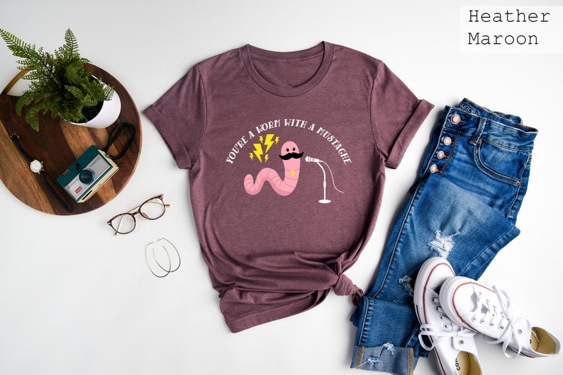 May include: A heather maroon t-shirt with a white graphic of a pink worm with a mustache holding a microphone. The text "You're a born with a mustache" is printed on the shirt.