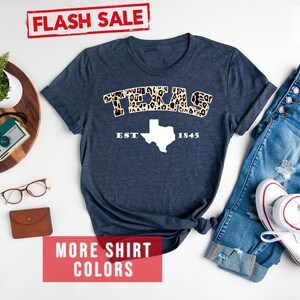 May include: A heather navy blue t-shirt with a white outline of the state of Texas and the word "TEXAS" in leopard print. The text "EST 1845" is below the state outline. The shirt is laid flat on a white surface with a pair of glasses, a camera, and a plant in a pot on a wooden tray to the left of the shirt. A pair of blue jeans and white trainers are to the right of the shirt. The text "FLASH SALE" is in a red rectangle at the top of the image. The text "MORE SHIRT COLORS" is in a red rectangle at the bottom of the image. The text "Heather Navy" is in black text at the top right of the image.