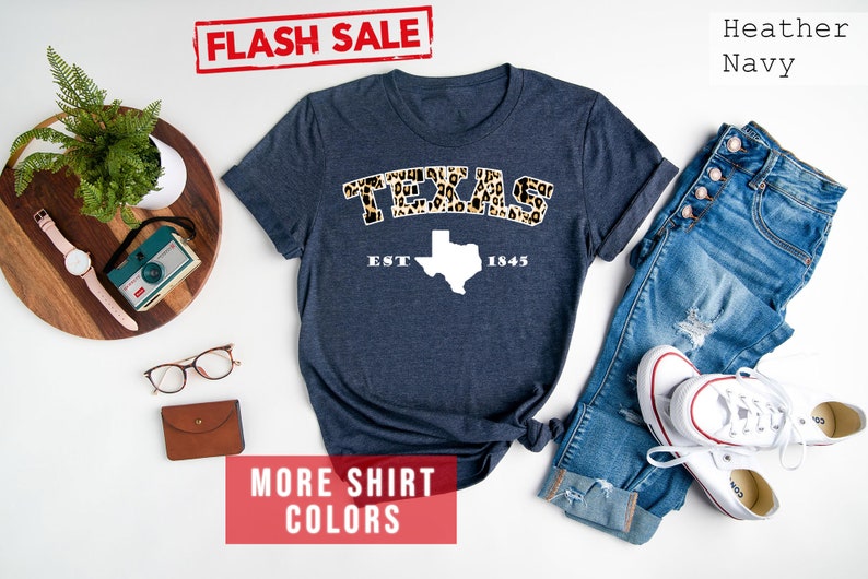 May include: A heather navy blue t-shirt with a white outline of the state of Texas and the word "TEXAS" in leopard print. The text "EST 1845" is below the state outline. The shirt is laid flat on a white surface with a pair of glasses, a camera, and a plant in a pot on a wooden tray to the left of the shirt. A pair of blue jeans and white trainers are to the right of the shirt. The text "FLASH SALE" is in a red rectangle at the top of the image. The text "MORE SHIRT COLORS" is in a red rectangle at the bottom of the image. The text "Heather Navy" is in black text at the top right of the image.