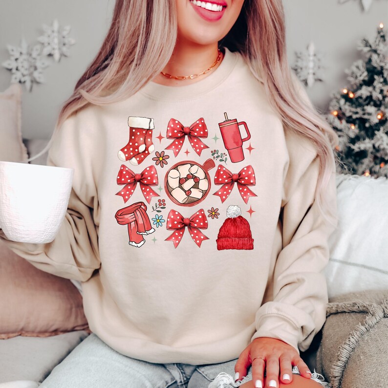 May include: Beige sweatshirt with a festive graphic featuring a red stocking, bows, scarf, mug, and a beanie. The design includes a cup of hot cocoa with marshmallows and floral accents. A cozy and seasonal apparel item.