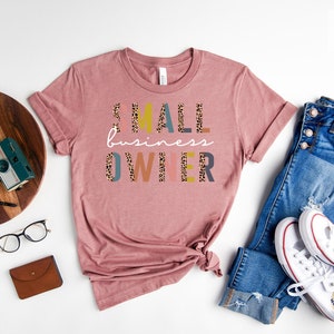 Pode incluir: Uma camiseta de cor malva desbotada com o texto "SMALL business OWNER" em letras coloridas com um padr&atilde;o de leopardo. A camiseta est&aacute; colocada sobre uma superf&iacute;cie branca com um par de cal&ccedil;as de ganga e t&eacute;nis brancos.