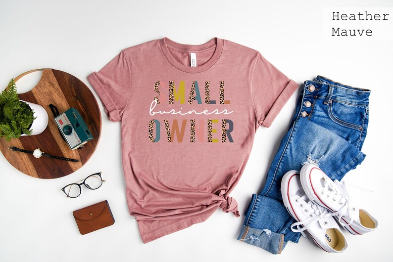 Pode incluir: Uma camiseta de cor malva desbotada com o texto "SMALL business OWNER" em letras coloridas com um padr&atilde;o de leopardo. A camiseta est&aacute; colocada sobre uma superf&iacute;cie branca com um par de cal&ccedil;as de ganga e t&eacute;nis brancos.