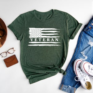 May include: A forest green t-shirt with a white distressed American flag and the word "Veteran" printed on it. The shirt is laid out on a white surface with a pair of glasses, a wallet, a camera, a plant, and a pair of jeans and white sneakers.