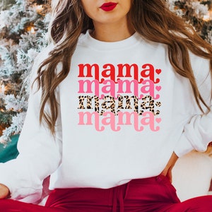 Op de afbeelding: Witte trui met de tekst "mama" die vier keer wordt herhaald in verschillende kleuren en stijlen. De tekst is in rood, roze, luipaardprint en roze met een hartje.