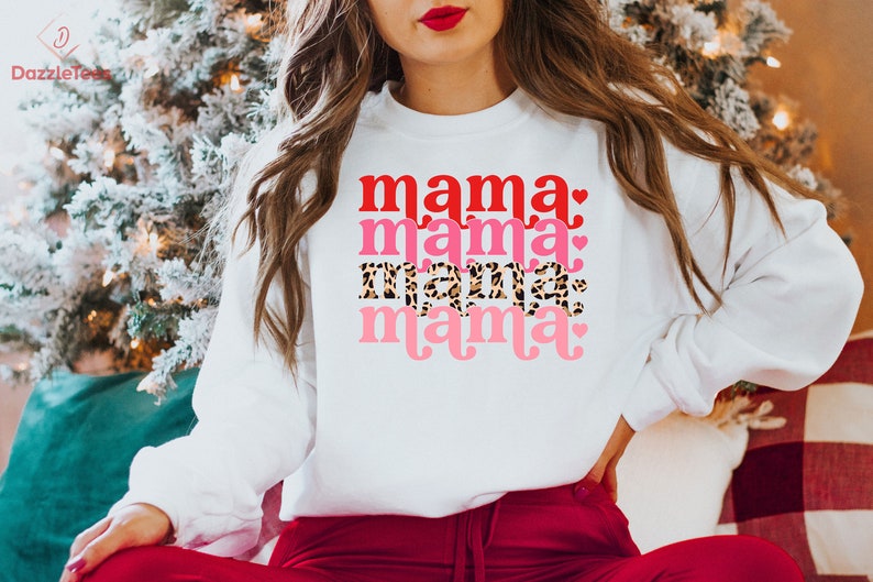 Op de afbeelding: Witte trui met de tekst "mama" die vier keer wordt herhaald in verschillende kleuren en stijlen. De tekst is in rood, roze, luipaardprint en roze met een hartje.