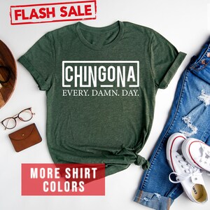 May include: A forest green t-shirt with the text "CHINGONA EVERY. DAMN. DAY." in white. The shirt is laid flat on a white surface with a pair of jeans, white sneakers, and other accessories.